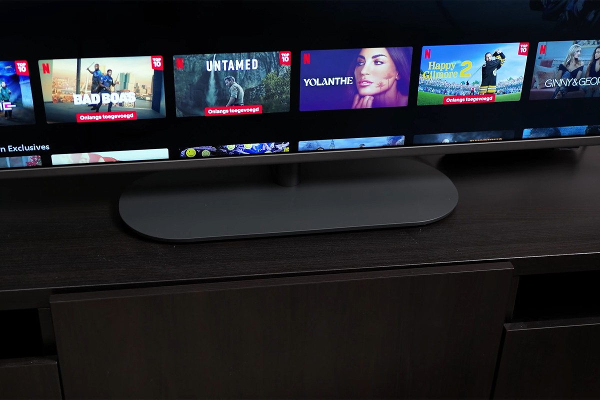 Draaibare standaard Philips The One 2025 Een televisie die op een donker tv-meubel staat, met op het scherm het Netflix-menu waarop diverse titels worden getoond zoals “BAD BOAS”, “UNTAMED”, “YOLANTHE”, “Happy Gilmore 2” en “GINNY & GEORGIA”, en met in het midden van het beeld de ovale voet van de tv goed zichtbaar