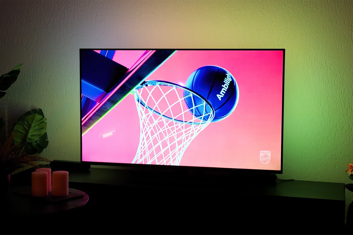 Philips The One Ambilight Philips The One 2025 waarop een kleurrijk beeld wordt weergegeven van een basketbal met het woord “Ambilight” die net door een basketbalring gaat, tegen een felroze achtergrond, terwijl de muur achter de tv verlicht wordt met groenige Ambilight-verlichting