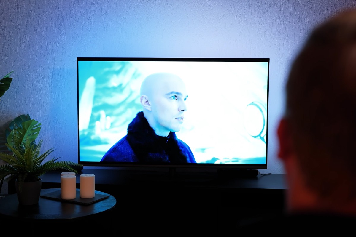 Philips The One 2025 Philips The One 2025 waarop een scène wordt afgespeeld van een kale man in een blauw getinte omgeving uit een film, terwijl een persoon op de voorgrond met zijn rug naar de camera toe zit te kijken; de muur achter de tv wordt blauw verlicht door Ambilight