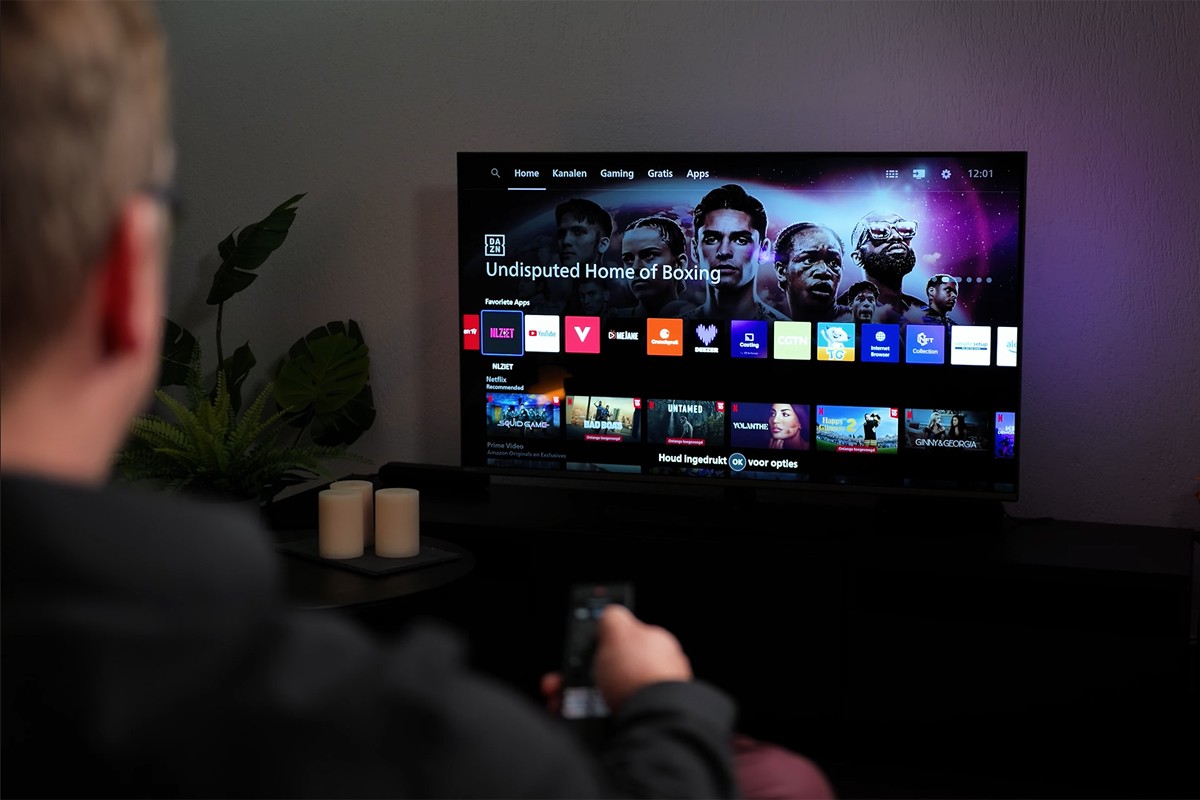 Philips The One 2025 Titan OS Een persoon zit op de bank en bedient met een afstandsbediening de Philips The One die het startscherm van Titan OS toont, met bovenaan het menu ‘Home’, ‘Kanalen’, ‘Gaming’, ‘Gratis’ en ‘Apps’, verder zijn apps zoals YouTube, NLZIET, Videoland en Netflix zichtbaar, en op de achtergrond is paarse Ambilight-verlichting te zien tegen de muur