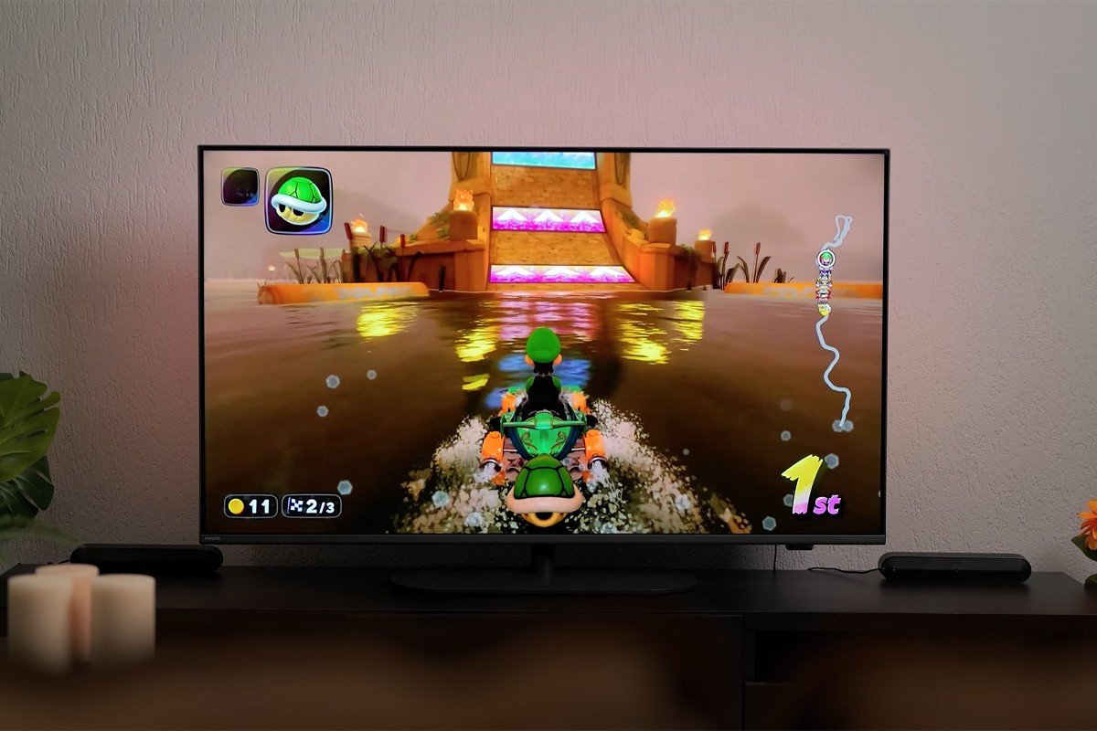 Philips The One 2025 gaming Philips The One 2025 waarop Mario Kart wordt gespeeld met personage Luigi dat op een wateroppervlak racet richting een tempelachtige helling met gekleurde lichtstroken; links onderin is te zien dat de speler 11 munten heeft en zich in ronde 2 van 3 bevindt, en rechts onderin staat dat hij op de eerste plaats ligt (“1st”), terwijl de muur subtiel verlicht wordt met Ambilight-verlichting.