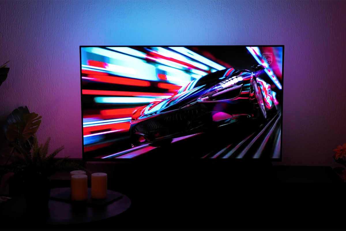 Philips The One 2025 Philips The One 2025 waarop een futuristisch ogende sportwagen met hoge snelheid door een tunnel van kleurrijke lichtstroken raast, met felle rode, blauwe en paarse tinten die via Ambilight-verlichting sfeervol op de muur achter de tv worden geprojecteerd
