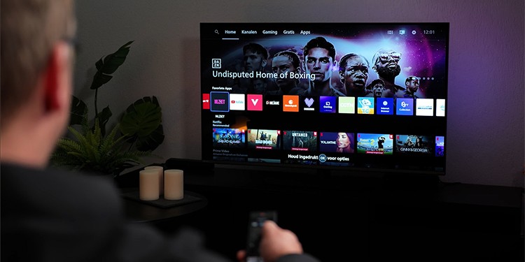 Een persoon zit op de bank en bedient met een afstandsbediening de Philips The One die het startscherm van Titan OS toont, met bovenaan het menu ‘Home’, ‘Kanalen’, ‘Gaming’, ‘Gratis’ en ‘Apps’, verder zijn apps zoals YouTube, NLZIET, Videoland en Netflix zichtbaar, en op de achtergrond is paarse Ambilight-verlichting te zien tegen de muur