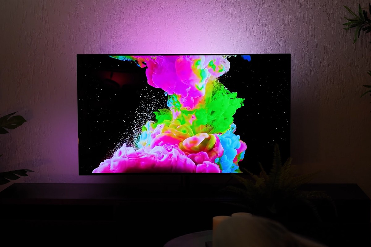 Philips OLED 2025 Philips OLED-tv die een explosie van kleurrijke vloeistoffen toont in felle tinten roze, groen, geel en blauw op een zwarte achtergrond terwijl de muur achter de tv wordt verlicht met paarse Ambilight-verlichting die de levendige kleuren op het scherm versterkt.