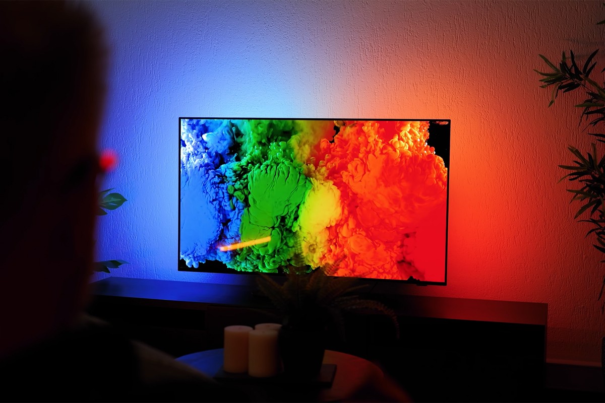 Philips OLED 2025 Philips OLED-tv toont een kleurrijke explosie van inktachtige wolken in blauw, groen, geel en rood op een zwarte achtergrond terwijl Ambilight-verlichting aan de muur.