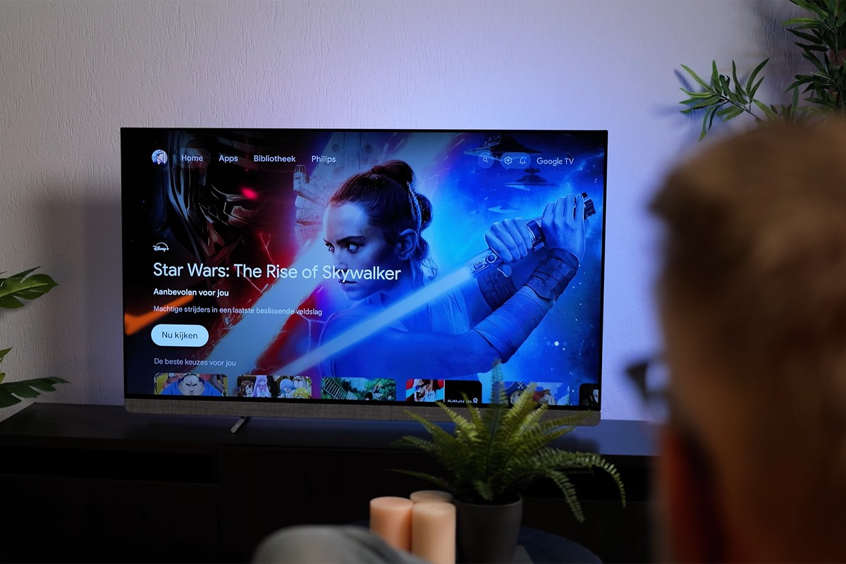 Philips OLED Philips OLED-tv waarop het startscherm van Google TV zichtbaar is met als uitgelichte titel “Star Wars: The Rise of Skywalker”. Een man kijkt naar het scherm, terwijl de muur op de achtergrond blauw verlicht is door Ambilight.
