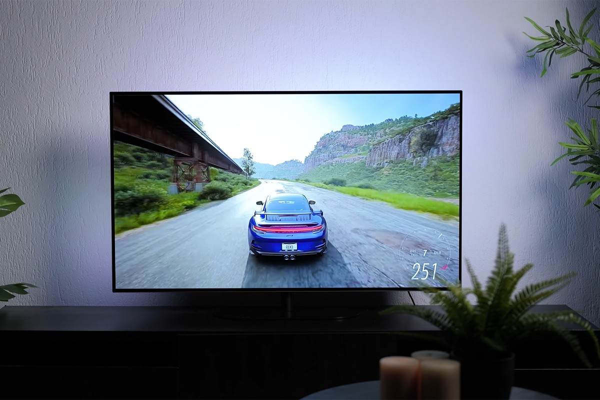 Philips OLED Philips OLED-tv waarop een racespel wordt gespeeld met een blauwe sportwagen die met hoge snelheid (251 km/u) over een weg rijdt langs rotswanden en een spoorbrug; het camerastandpunt is achter de auto, en de muur achter de tv is subtiel blauw verlicht door Ambilight-verlichting.