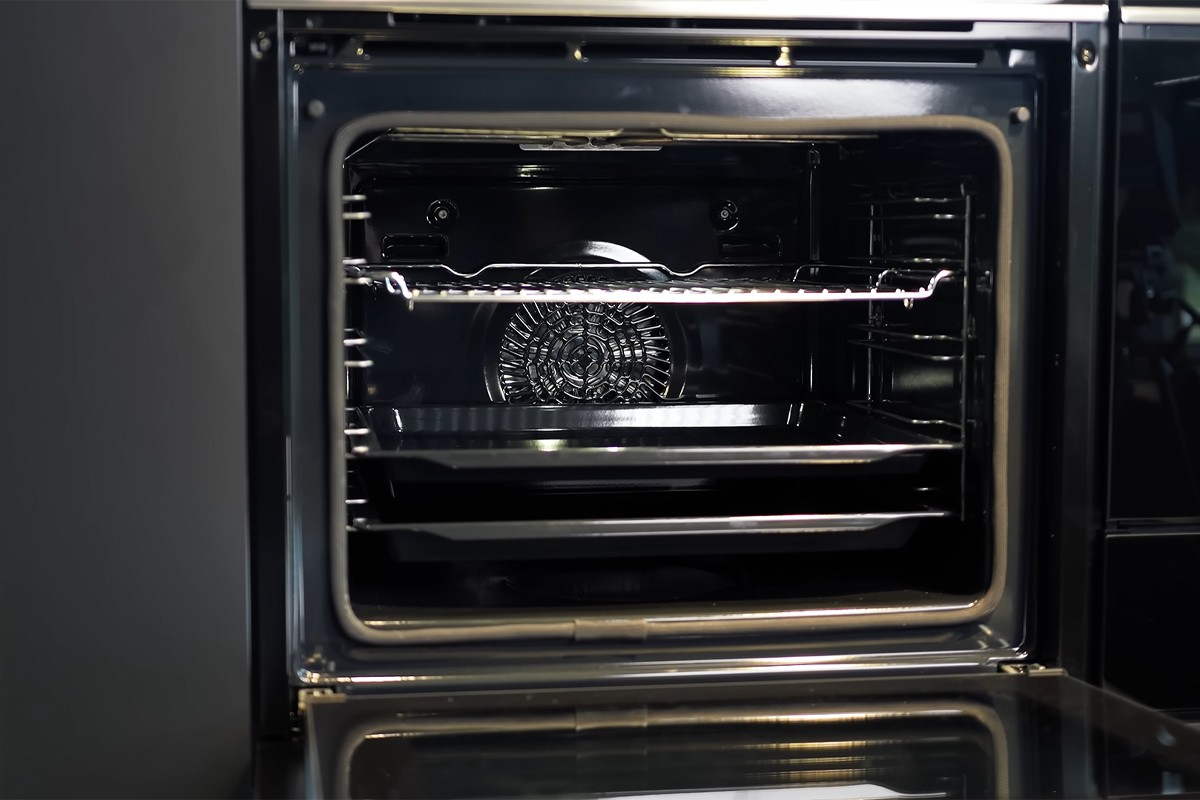 Een open inbouwoven met een schone, glanzende binnenkant; zichtbaar zijn een metalen rooster in het midden en een bakplaat eronder, met achterin de oven een ventilator voor heteluchtfunctie.