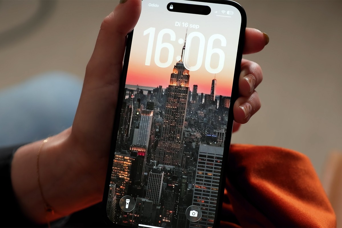 Een persoon houdt een iPhone vast met het vergrendelscherm actief, waarop de tijd 16:06 wordt weergegeven op dinsdag 16 september, en op de achtergrond een spectaculaire foto van de skyline van New York met de Empire State Building prominent in beeld tegen een oranje-roze zonsondergang.