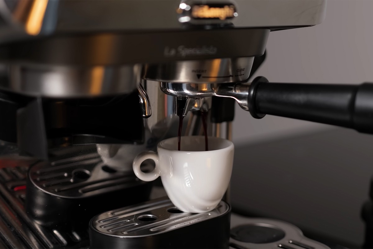 Close-up van een De'Longhi La Specialista Touch espressomachine tijdens het zetten van koffie; er stroomt verse espresso uit de dubbele uitloop van de filterdrager rechtstreeks in een klein wit espressokopje