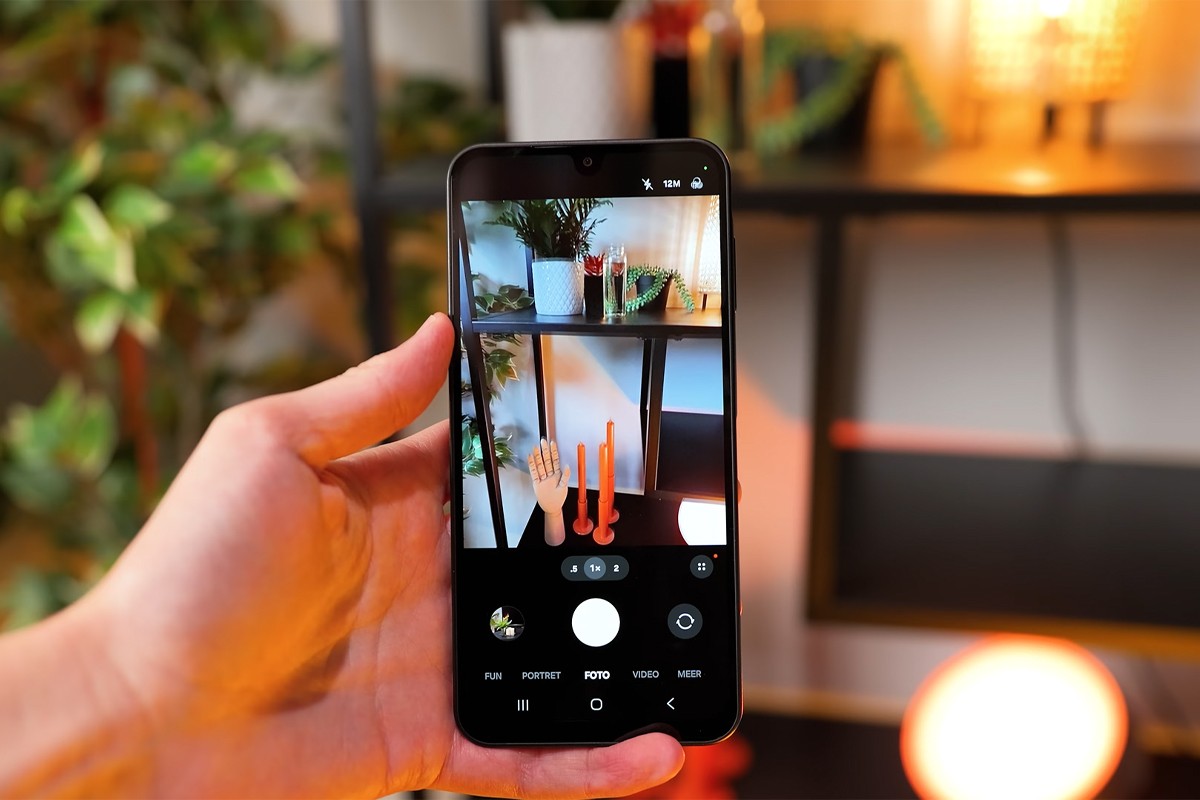 Samsung Galaxy A17 Een hand houdt een Samsung Galaxy A17-smartphone vast met de camera-app geopend in de fotomodus; op het scherm wordt een kleurrijk interieur vastgelegd met planten, rode kaarsen en decoratieve objecten op een metalen rek
