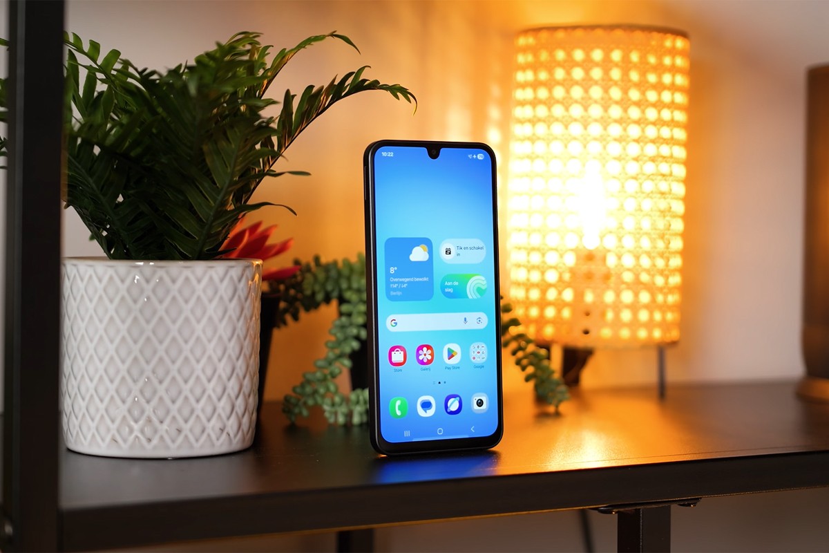 Samsung Galaxy A17 Samsung Galaxy A17 staat rechtop op een plank naast een witte plantenpot en een sfeervolle tafellamp