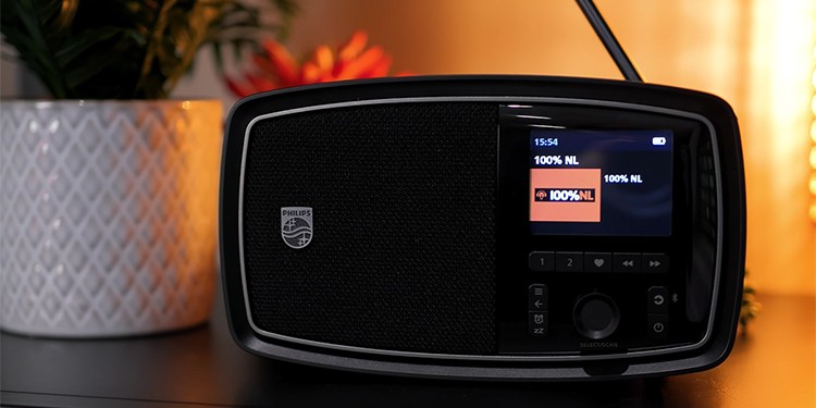 Philips The Janet radio met het station “100% NL” actief op het scherm. De radio heeft een zwart ontwerp met een display aan de rechterkant en een luidspreker aan de linkerkant.