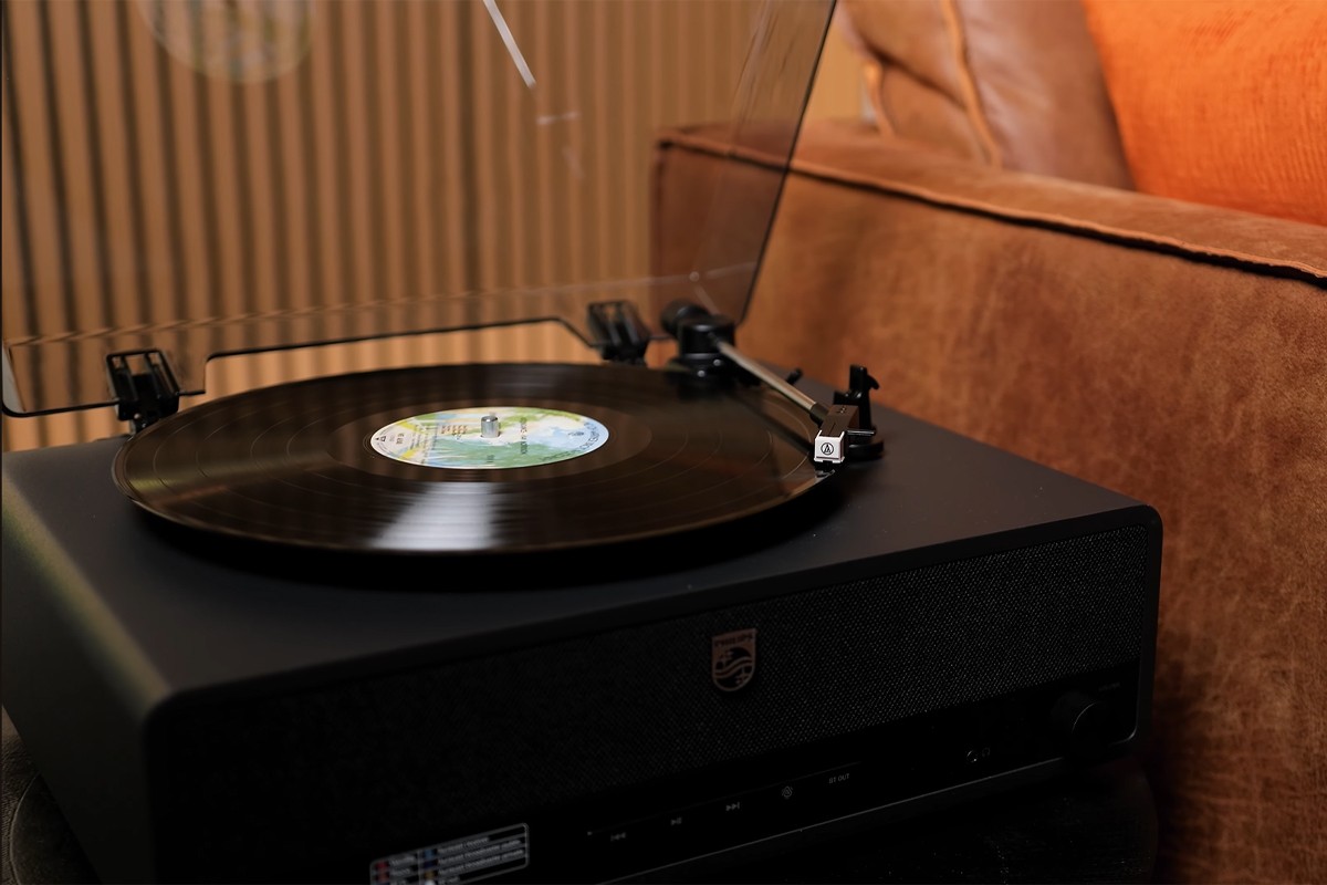 Philips The Stevie Philips The Stevie met een transparante stofkap, waarop een vinylplaat draait, geplaatst naast een bruine leren bank.