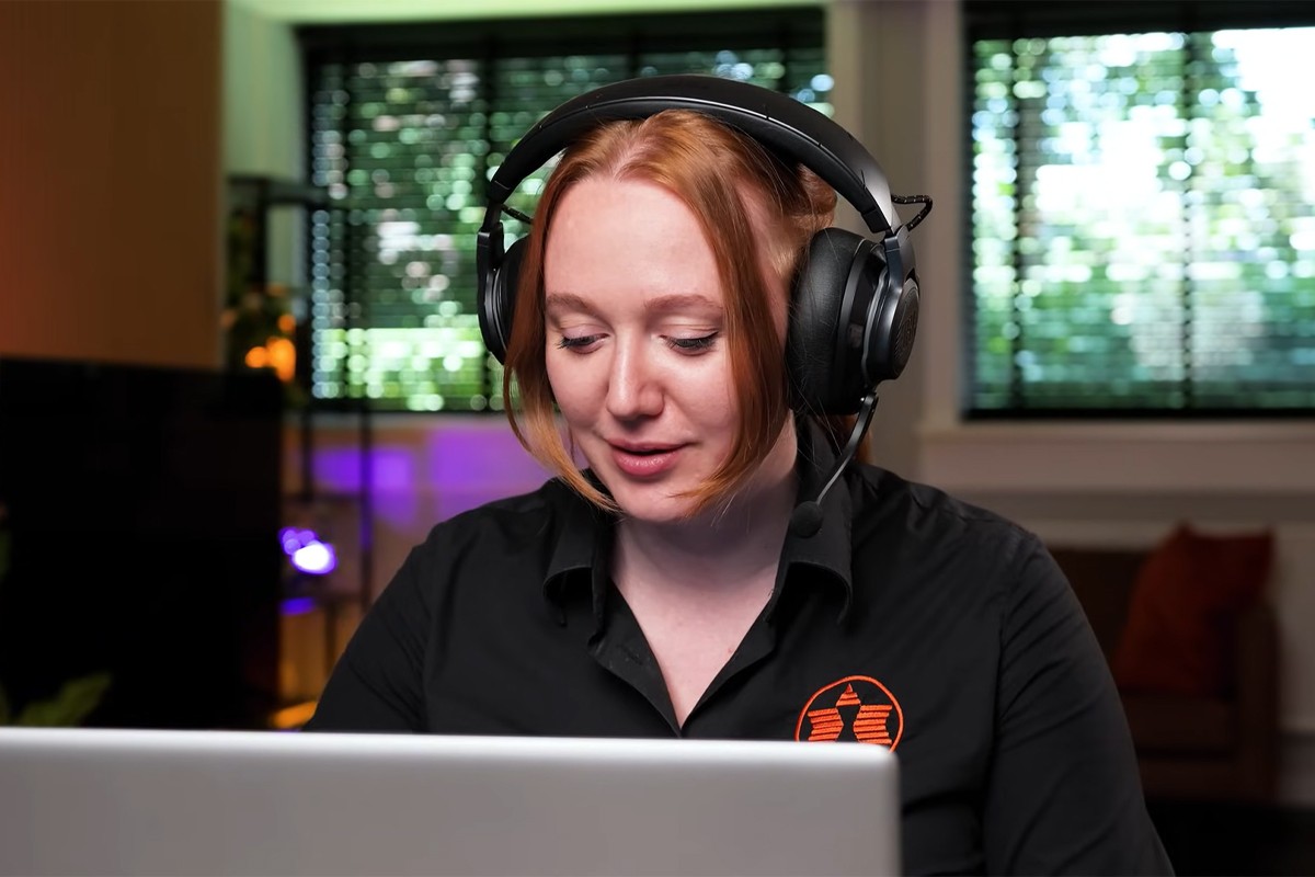 Een vrouw draagt een grote, zwarte gamingheadset met microfoon terwijl ze achter een opengeklapte laptop zit. Ze draagt een zwart overhemd met Expert-logo.