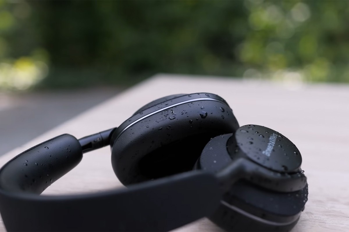 Een close-up van een zwarte over-ear koptelefoon die plat ligt op een houten oppervlak, met zichtbare waterdruppels op de oorschelpen en hoofdband.