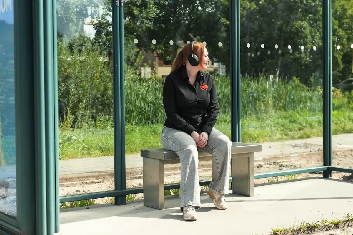 Een vrouw met een zwarte over-ear koptelefoon zit op een metalen bankje in een bushokje.