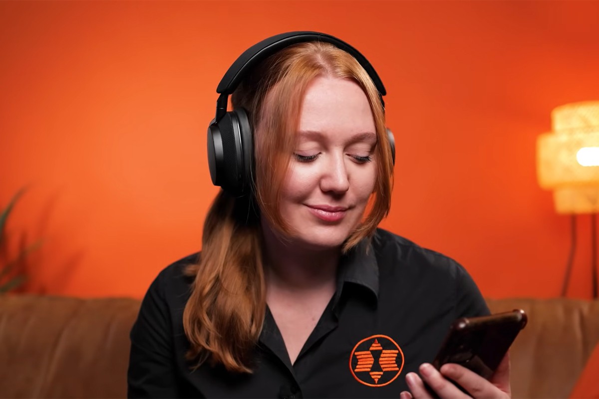 Een vrouw draagt een zwarte over-ear koptelefoon en kijkt ontspannen naar haar smartphone. Ze zit op een bank met een feloranje muur op de achtergrond. Ze draagt een zwarte blouse met een oranje Expert-logo.