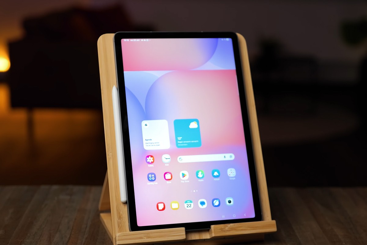 Close-up van de Samsung Galaxy Tab S10 Lite op een houten standaard, met het scherm aan en verschillende app-iconen zichtbaar; de tablet is geflankeerd door een stylus aan de zijkant.