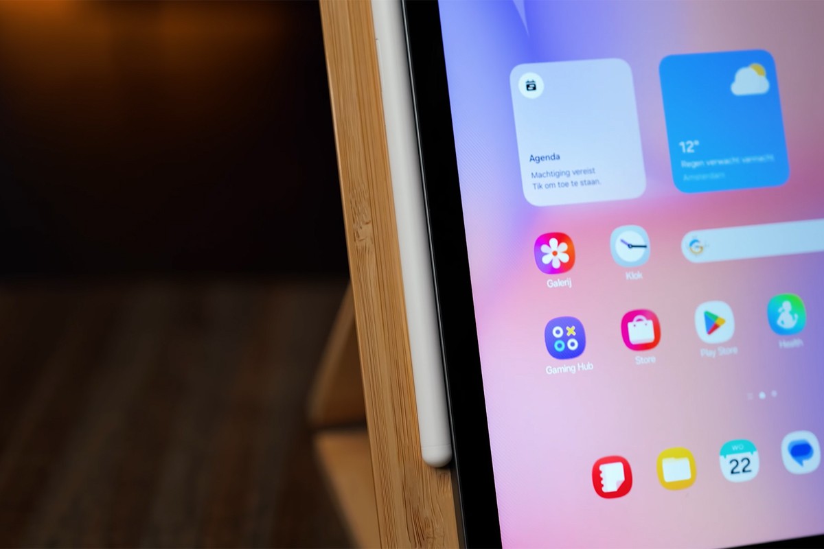 Close-up van de Samsung Galaxy Tab S10 Lite op een houten standaard, met de witte S Pen netjes magnetisch bevestigd aan de zijkant van het scherm.