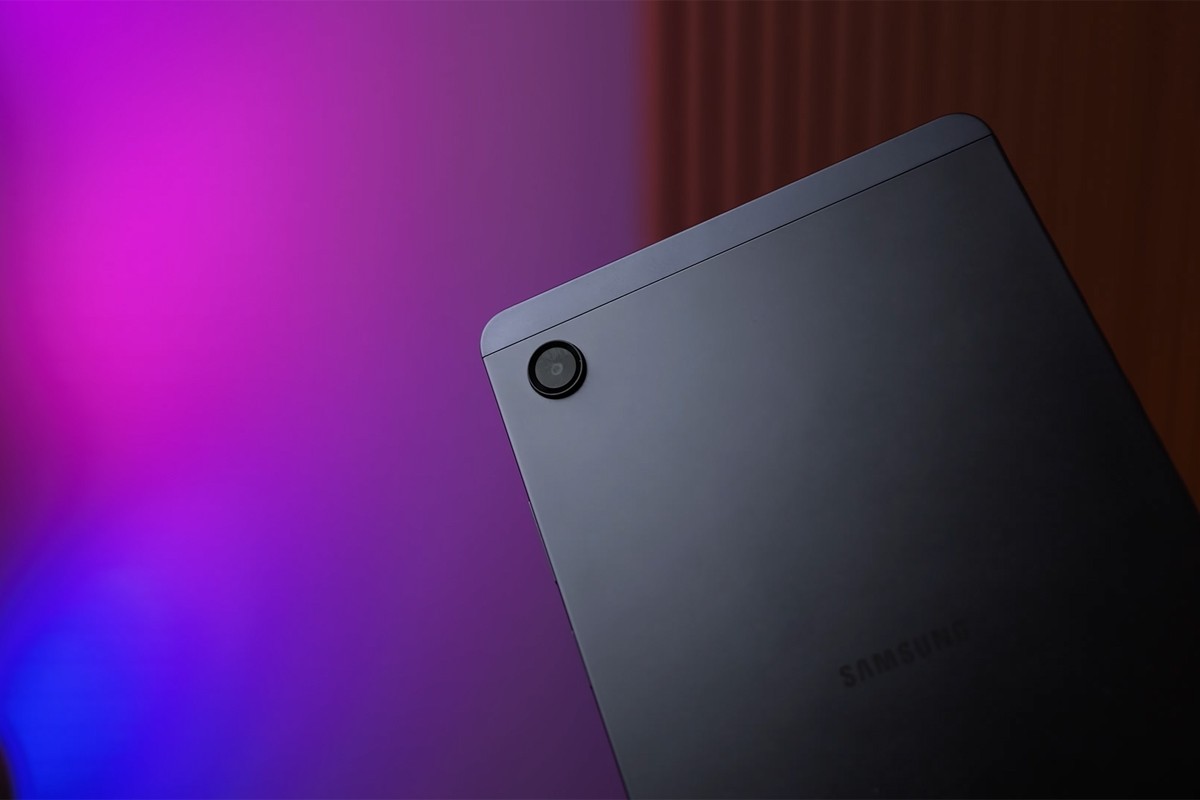 Close-up van de achterkant van een Samsung Galaxy Tab A11-tablet; de tablet is donkergrijs van kleur en staat schuin in beeld tegen een kleurrijke achtergrond met paarse, roze en blauwe verlichting.