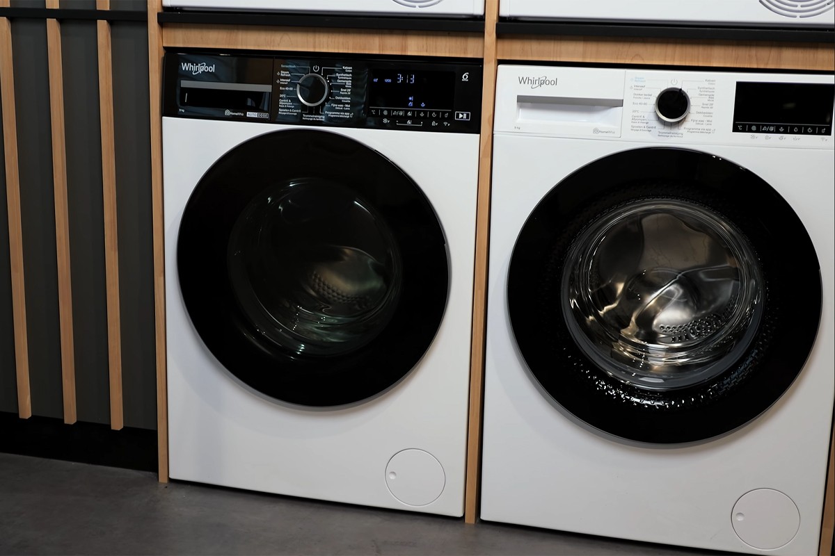 Close-up van twee Whirlpool-wasmachines die naast elkaar in een showroomopstelling staan; links is een model te zien met een zwart glanzend bedieningspaneel en een draaiknop met programma-indeling rondom, rechts een vergelijkbaar model met een eenvoudiger wit paneel en een draaiknop met een lijst aan programma’s eromheen.