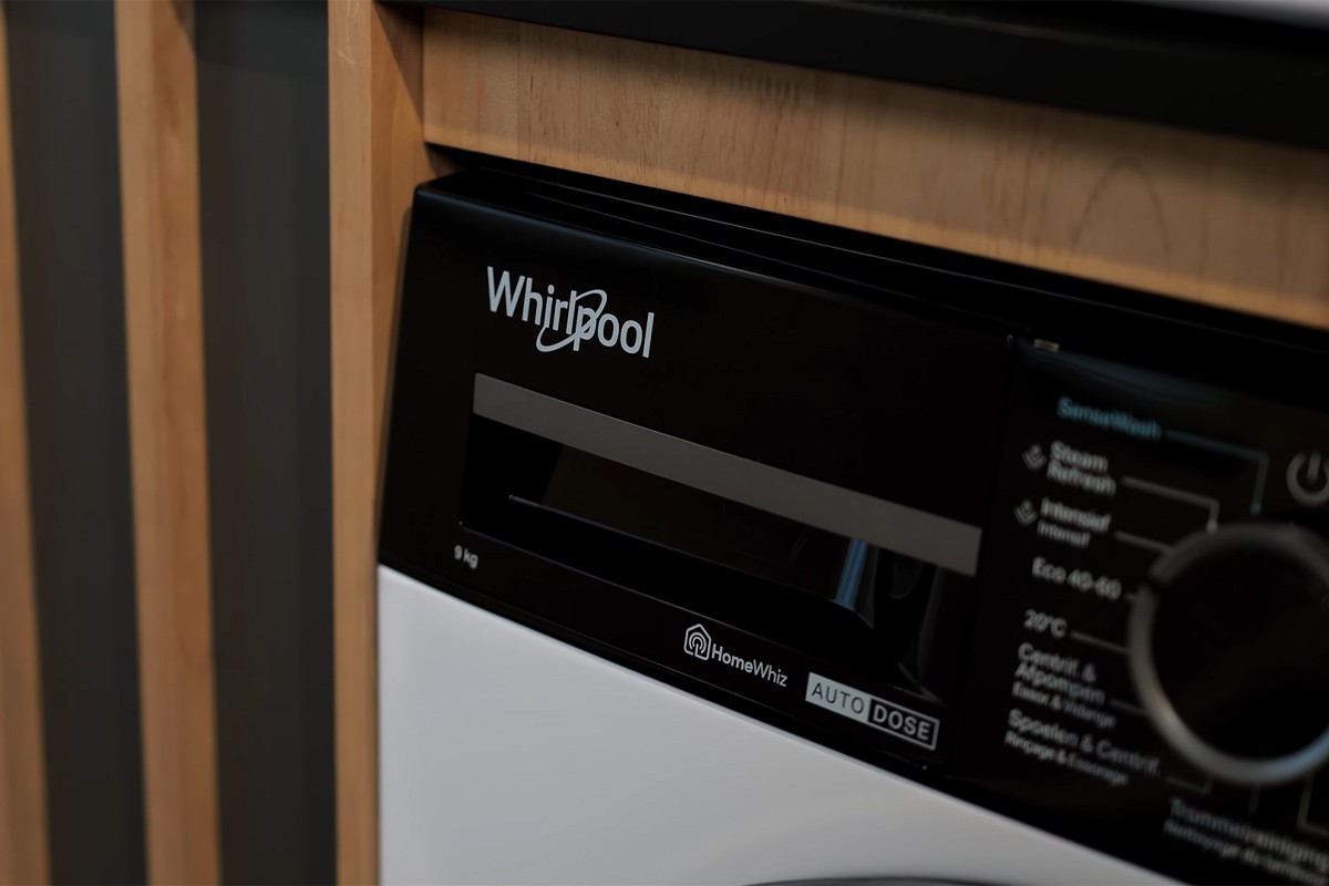 Close-up van het bedieningspaneel van een Whirlpool-wasmachine, ingebouwd in een houten ombouw. Linksboven is het Whirlpool-logo zichtbaar op een zwart glanzend oppervlak, daaronder een gleuf voor wasmiddeltoevoer, met daarnaast aanduidingen zoals “HomeWhiz” en “AUTO DOSE”.