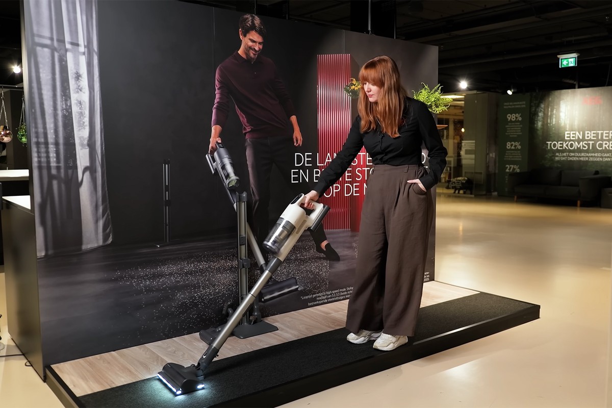 Een vrouw demonstreert een steelstofzuiger in een moderne winkel, met een grote productposter op de achtergrond.