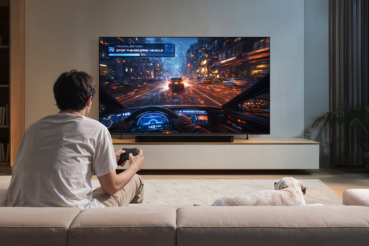 Persoon zit op een beige kleuige bank met een controller van een gameconsole in zijn handen. Rechts naast hem ligt een hond en beide kijken ze naar het scherm van de Samsung M80H waarop een AI-gegenereerde gameplay te zien is van een racespel.