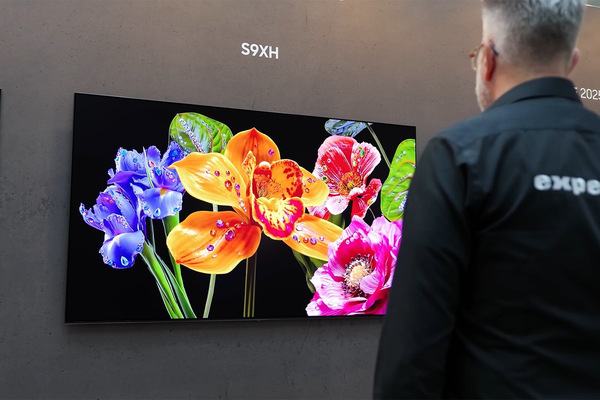 Presentator met Expert-blouse kijkt naar de Samsung OLED S93H aan de muur.