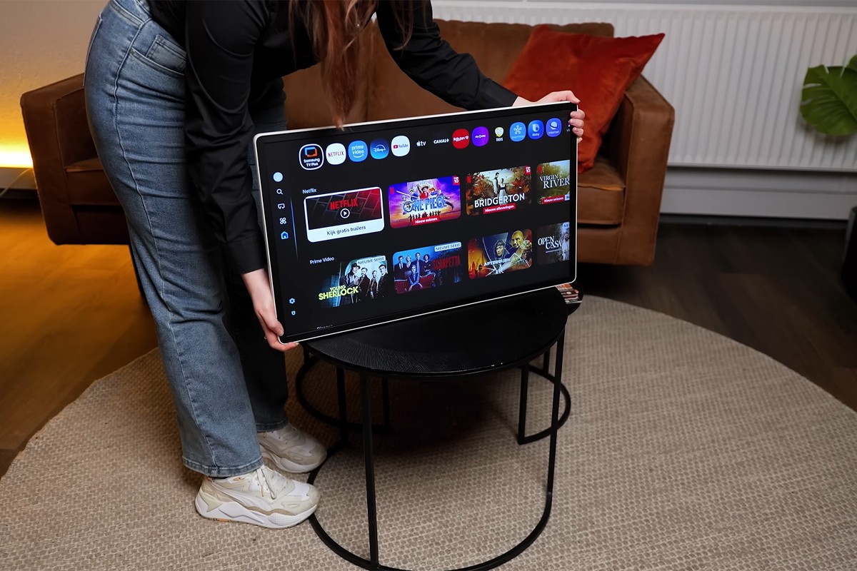 Scherm van de Samsung The Movingstyle losgekoppeld van het standaard en wordt door een persoon op een tafel in de woonkamer gezet. Op het scherm is het Tizen OS besturingssysteem duidelijk zichtbaar.
